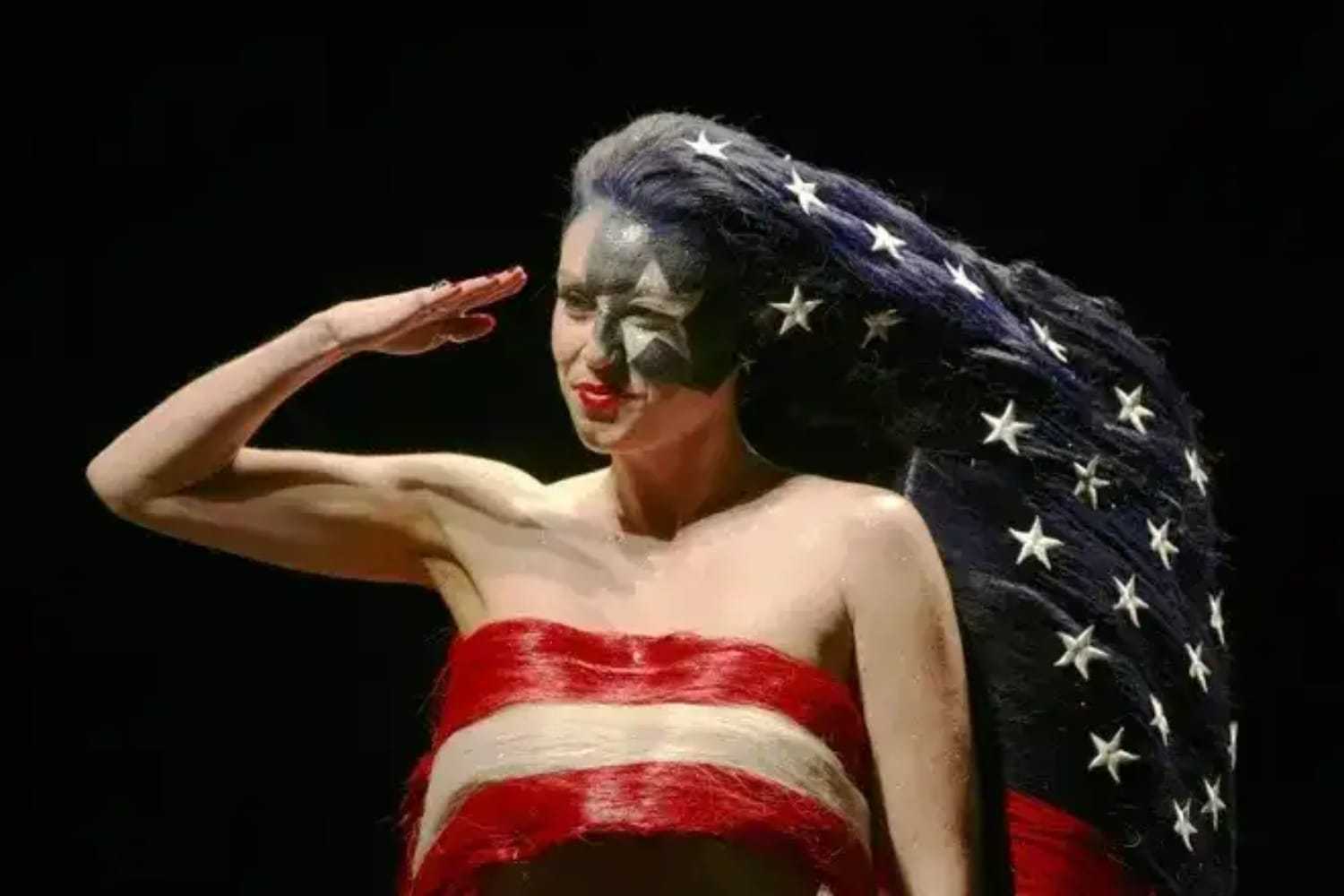 A woman salutes with hair painted as the American flag, featuring stars and stripes.